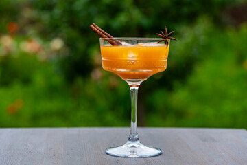 Healthy pumpkin fruit smoothie with ice decorated with star anise and cinnamon stick in elegant crystal glass on wooden table against of a green summer garden, closeup