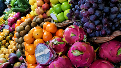A vibrant fruit stall features a rich selection of tropical produce displayed in woven baskets. Bananas, avocados, pineapples, mangoes, pomegranates, and a distinctive green fruit