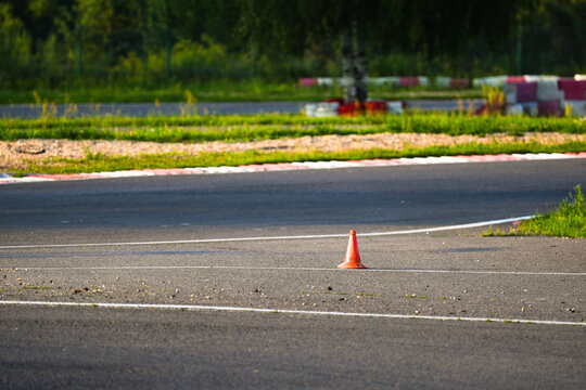 Single orange traffic cone placed at the curve of an asphalt go-kart track in bright sunlight. Green grass, red-white curbs, and blurred fencing in the background. Ideal image for safety, motorsport