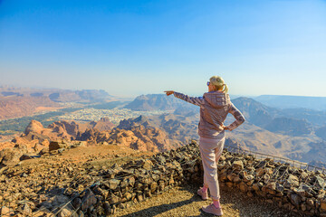 Blonde tourist pointing at AlUla Old Town from the Harrat Viewpoint, enjoying breathtaking panoramic views of the Saudi Arabian desert landscape in Al-Ula, Madinah province
