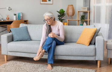 Sad caucasian elderly gray-haired woman in glasses sit on sofa touches leg, suffering from paine in...