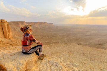 Female tourist wearing hat and sunglasses sitting on rock admiring sunset view of Tuwaiq...