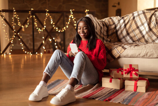 Merry Christmas. Happy black woman in warm sweater using smartphone, shopping online, choosing gifts, sitting on floor near boxes in decorated living room. Winter holiday season concept