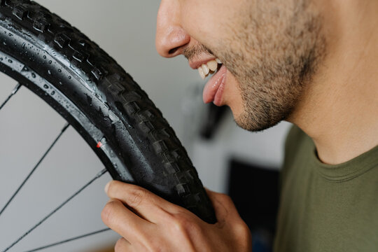 Close-up cropped shot cheerful man licking tire of mountain bike wheel while holding rag, showcasing humorous approach to bicycle maintenance, side view portrait. Concept of bicycle transport service.