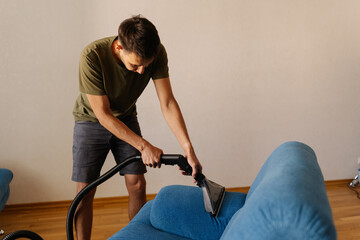 Side view of focused man in causal clothing using washing vacuum cleaner to clean blue sofa in home...