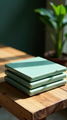 Three green notebooks stacked on a wooden table near a green plant, suggesting study or work.