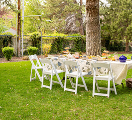 An outdoor tea party table set up in a garden with white folding chairs, floral tablecloth, snacks,...