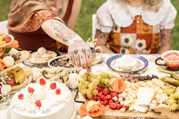 tattooed hand reaching for grapes on a floral tablecloth-covered outdoor picnic table filled with a cheese board, desserts, fruit, and vintage tea ware