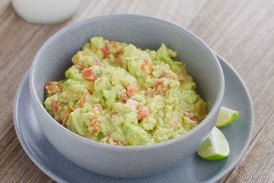 Freshly prepared homemade guacamole dip or salsa, made of avocado, tomato, lime juice, salt and pepper, served in blue bowl with lime wedges on the side (Selective Focus, Focus in the middle of the im
