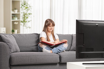 Little girl sitting on a sofa and reading a book in front of tv