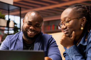 Young adults in their living space browsing webpages on a laptop together while they sit on the...