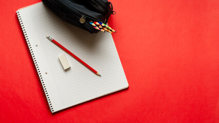 A notebook with a pencil and an eraser, and a pencil case full of colorful pencils on red background and copy space. Back to school concept. 
