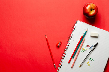 An apple, a notebook, some colorful pencils, a pen, an eraser, a sharpener, and paperclips on red background and copy space. Back to school concept. 