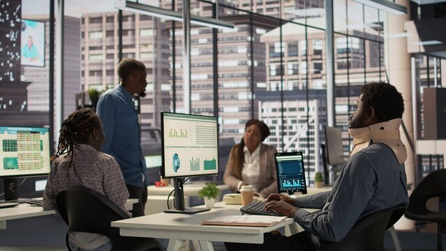 Black businessman struggles to work at the desk with a neck fracture, dealing with pain while he wears cervical collar for protection and support. Injured worker tries to solve tasks. Camera B.