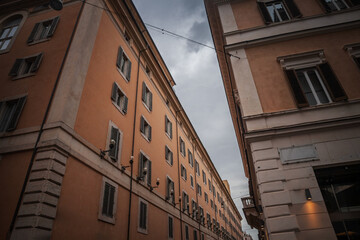 Looking up a narrow Roman street, warm apartment blocks with shuttered windows converge on a brooding sky, lamps and weathered masonry framing the residential face of Italy's capital in rome italy.