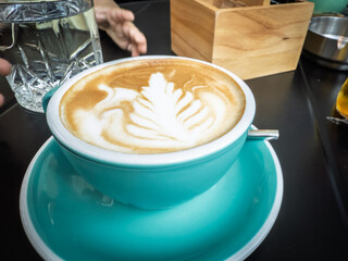 Turquoise cup filled with creamy cappuccino topped by leaf‑shaped latte art sits on a matching saucer at a modern cafe table, crystal water glass and blurred hands visible in the background.