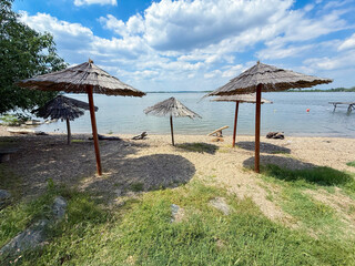 Weathered reed parasols cast thin shadows across a narrow pebble beach on the Danube near Belgrade,...