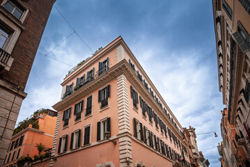 Low angle perspective of a pastel salmon Roman apartment building with shuttered windows, trailing rooftop greenery and quoins, rising sharply between narrow streets under a clear morning sky.