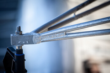 Closeup view of a clean TIG weld seam joining two silver metal rods in a lightweight frame. Shallow depth of field isolates the shiny bead, bolt and nut while surrounding workshop fades to gray blur.
