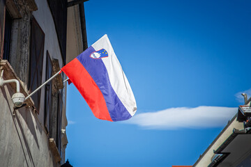 The red, blue and white flag of Slovenia with national shield ripples from a wall mounted pole against a deep blue sky and small fair weather cloud, captured in bright midday light in a coastal town.