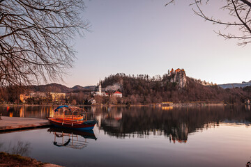Twilight settles over Lake Bled in Slovenia as a wooden pletna boat rests at the jetty, mirroring on glassy water beneath Bled Castle and the spire of St Martin's Church rising from the shoreline.