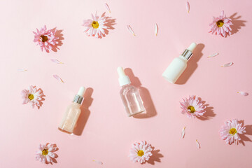 Cosmetic serum bottles on light pink background with pink daisy flowers, petals and hard shadows....