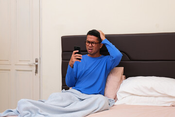 Adult Asian man on bed showing confused expression when looking at his phone