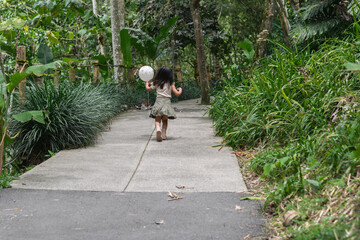 Little Latina girl strolls down concrete zoo path holding a white balloon amid tropical plants.