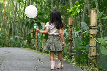 Latina toddler turns back clutching white balloon on bamboo-lined zoo path through lush forest.