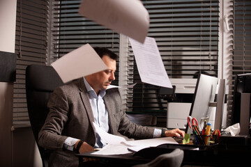 Stressed office worker typing on laptop as papers fly around him. Overwhelmed business atmosphere,...