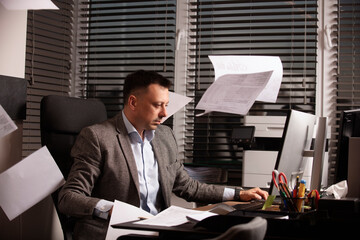 Stressed office worker typing on laptop as papers fly around him. Overwhelmed business atmosphere,...
