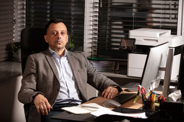 Pensive businessman in a blazer sitting at desk in modern office, staring thoughtfully at computer...