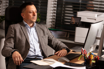 Pensive businessman in a blazer sitting at desk in modern office, staring thoughtfully at computer...