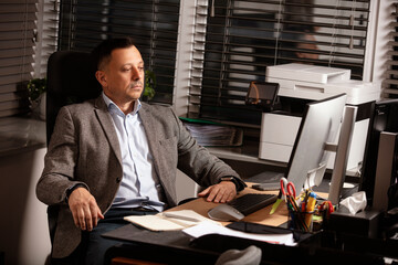 Pensive businessman in a blazer sitting at desk in modern office, staring thoughtfully at computer...