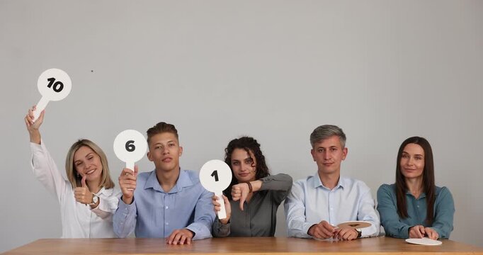 Panel of emotional judges voting with score signs at table near grey wall