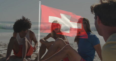 Sitting friends chatting in swimwear on sandy beach near shoreline, with Swiss flag and sunglasses