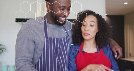 Couple hugging and cooking in home kitchen, with striped apron, red apron and hexagonal overlay
