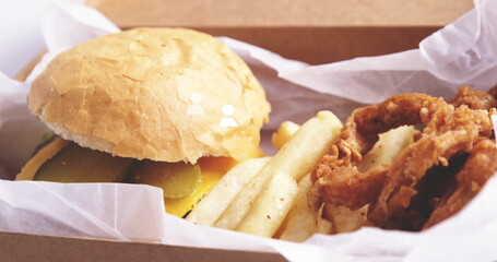 Takeout meal resting in box on table, with cheeseburger, pickles, fries, onion rings, copy space