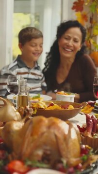 Smiling woman and boy sitting at festive table with roasted turkey, autumn leaves, wine glasses, and traditional food in daylight interior. Indoor lifestyle photography. Thanksgiving celebration. Fami