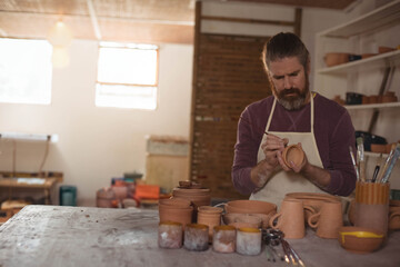 Mid adult male potter wearing apron applying glaze on clay pot at workshop workbench, copy space