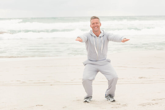 Mature man squatting with arms extended wearing light gray hoodie and sweatpants on sandy shoreline