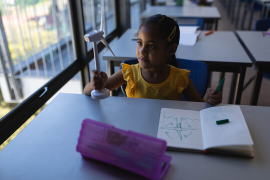 Student is holding small wind turbine model and grasping green marker by notebook on classroom desk