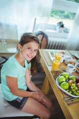 Cheerful preteen girl sitting at breakfast table beside window with green apples, grapes and juice