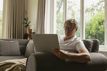 Senior man sitting on gray sofa in home living room using laptop next to potted plant