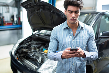Man wearing striped shirt standing in auto repair shop holding smartphone near car with open hood