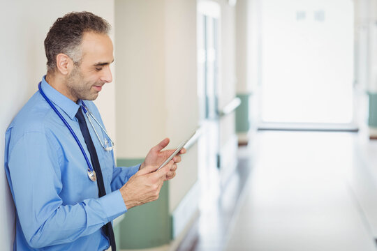 Middle-aged male doctor leaning on wall using tablet in hospital hall with stethoscope, copy space - Powered by Adobe