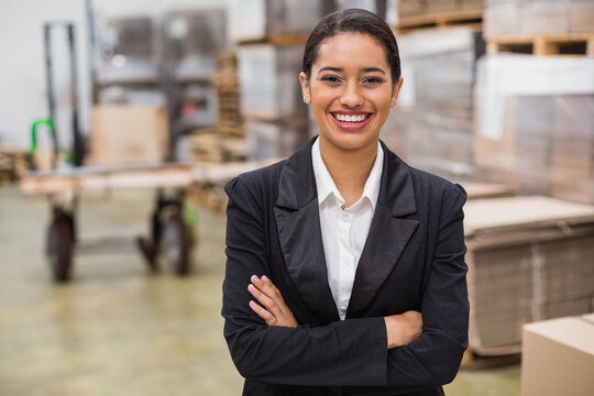 Hispanic woman standing with crossed arms and smiling in warehouse with pallet jack and boxes