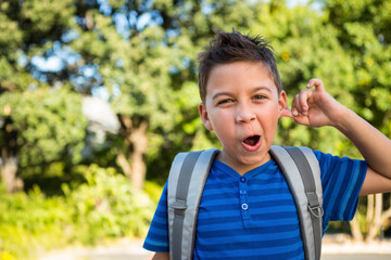 Boy pulling ear and making playful face while standing outdoors in park with backpack, copy space