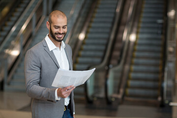 Man standing in large lobby reading folded newspaper near parallel escalators with polished floor