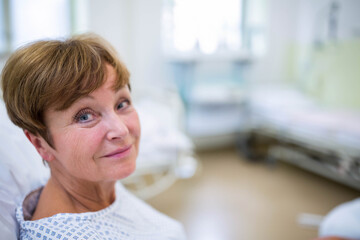 Senior woman reclining on hospital bed near empty bed in patient room wearing patterned gown